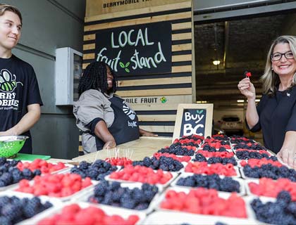 Une femme tient une fraise devant un présentoir de fruits locaux.