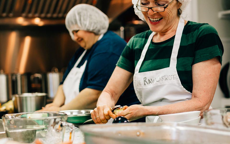 Two women wearing aprons peeling carrots in a kitchen.