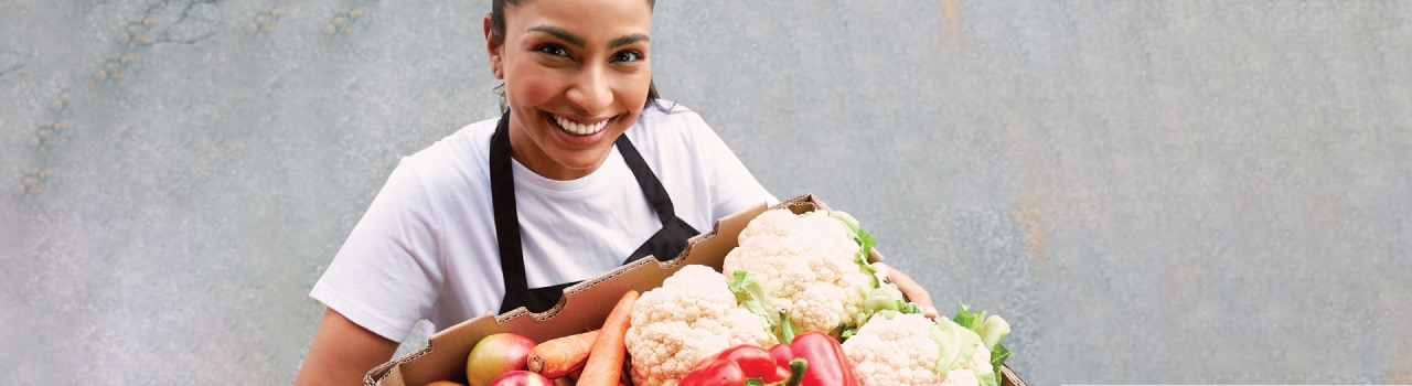 This banner image shows a woman holding a fresh vegetable box and smiling while getting her picture taken.