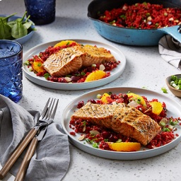 Two plates of grilled salmon with quinoa and orange slices on a white table with blue glasses, utensils, and a pan of more food in the background.